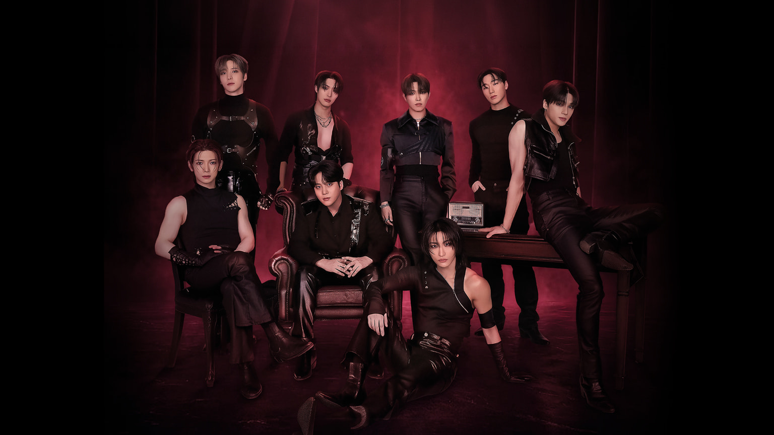 "Gruppenfoto der K-Pop-Band ATEEZ in schwarzen Lederoutfits vor rotem, rauchigem Bühnenvorhang, teils sitzend, teils stehend in dramatischer Pose"